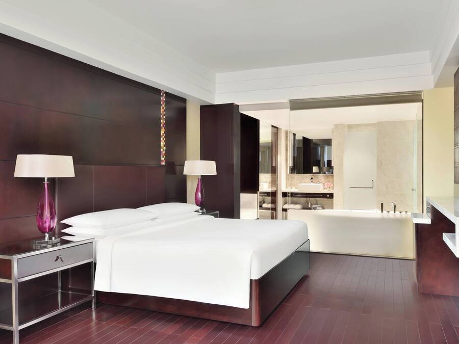 Una habitación de hotel moderna con una cama grande, dos mesitas de noche con lámparas, una bañera empotrada y un espejo amplio.