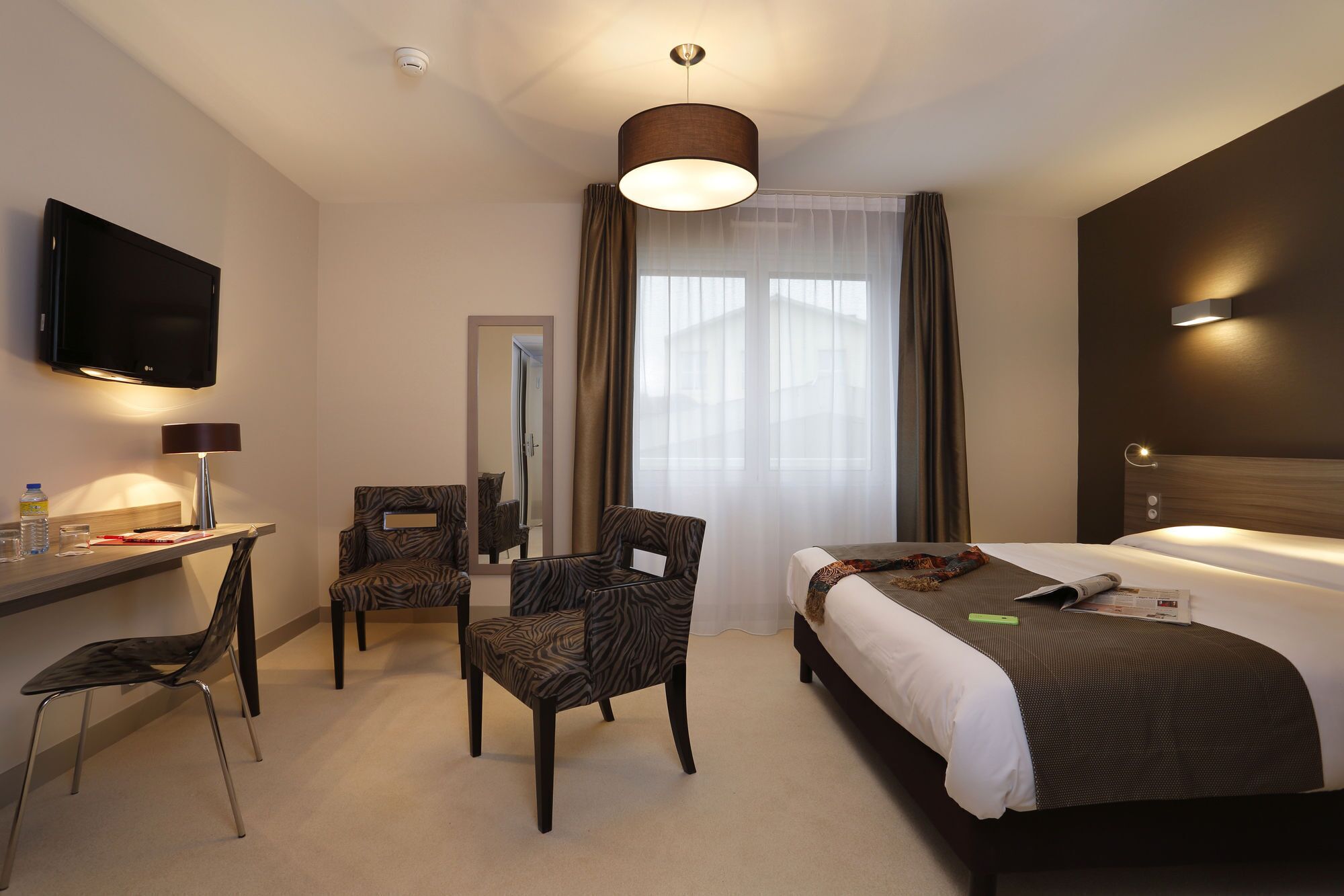 A hotel room with a bed, a desk with a chair, a TV, and a large window with curtains.
