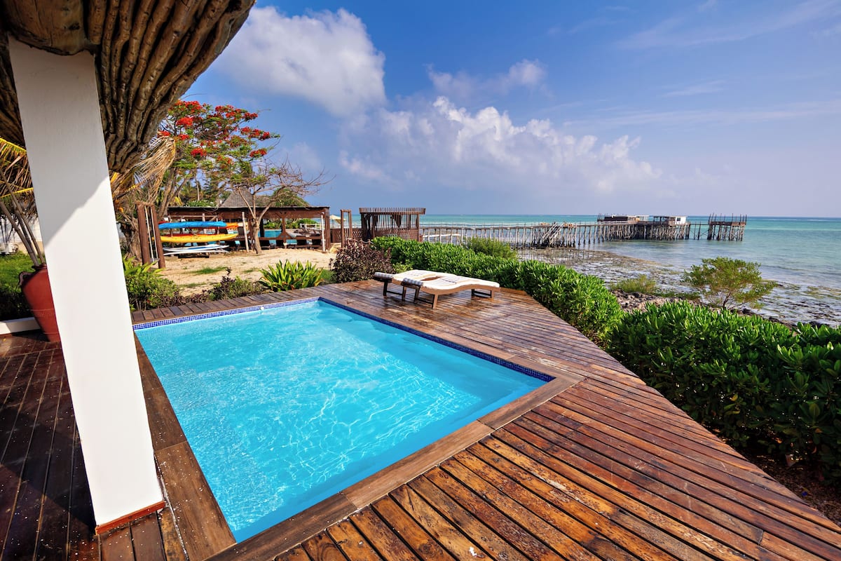 A wooden deck with a swimming pool, lounge chairs, and a view of the ocean and pier.