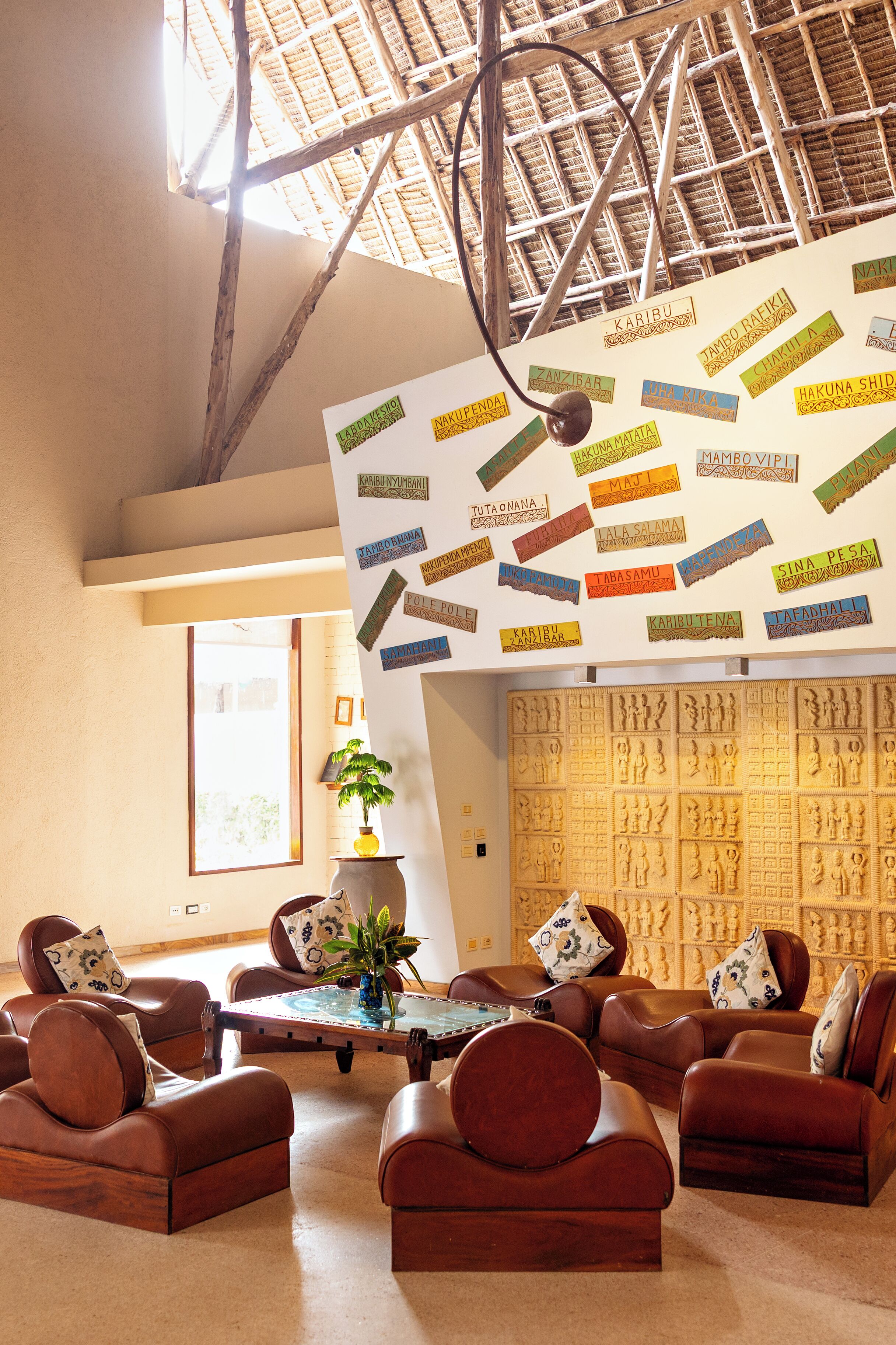 Ein Zimmer mit hohen Decken, einem Strohdach und einer Wand mit geschnitzten Paneelen. Mehrere Ledersofas sind um einen zentralen Couchtisch angeordnet.