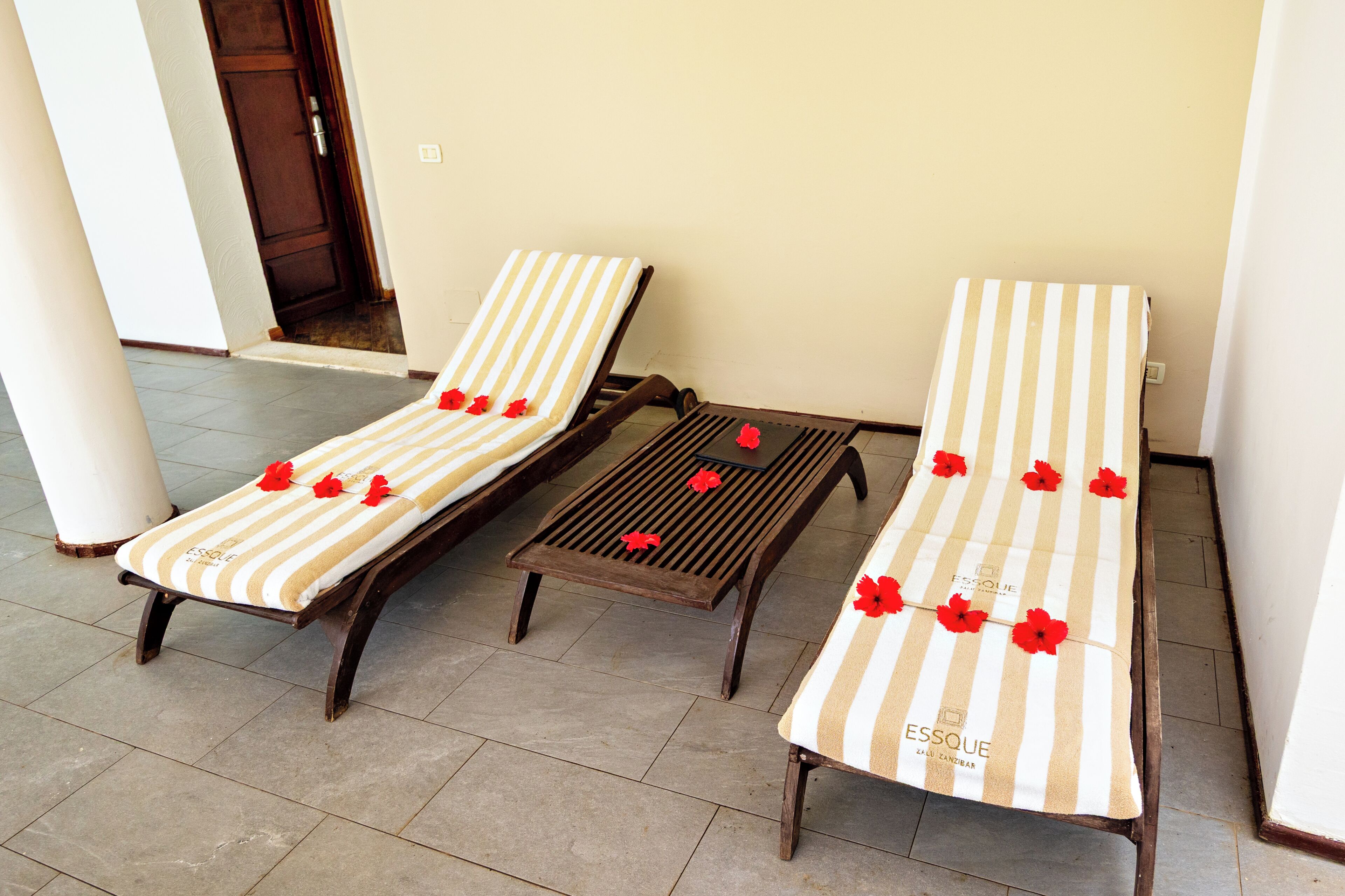 Two sun loungers with striped cushions and red flowers, a wooden table, and a beige wall.