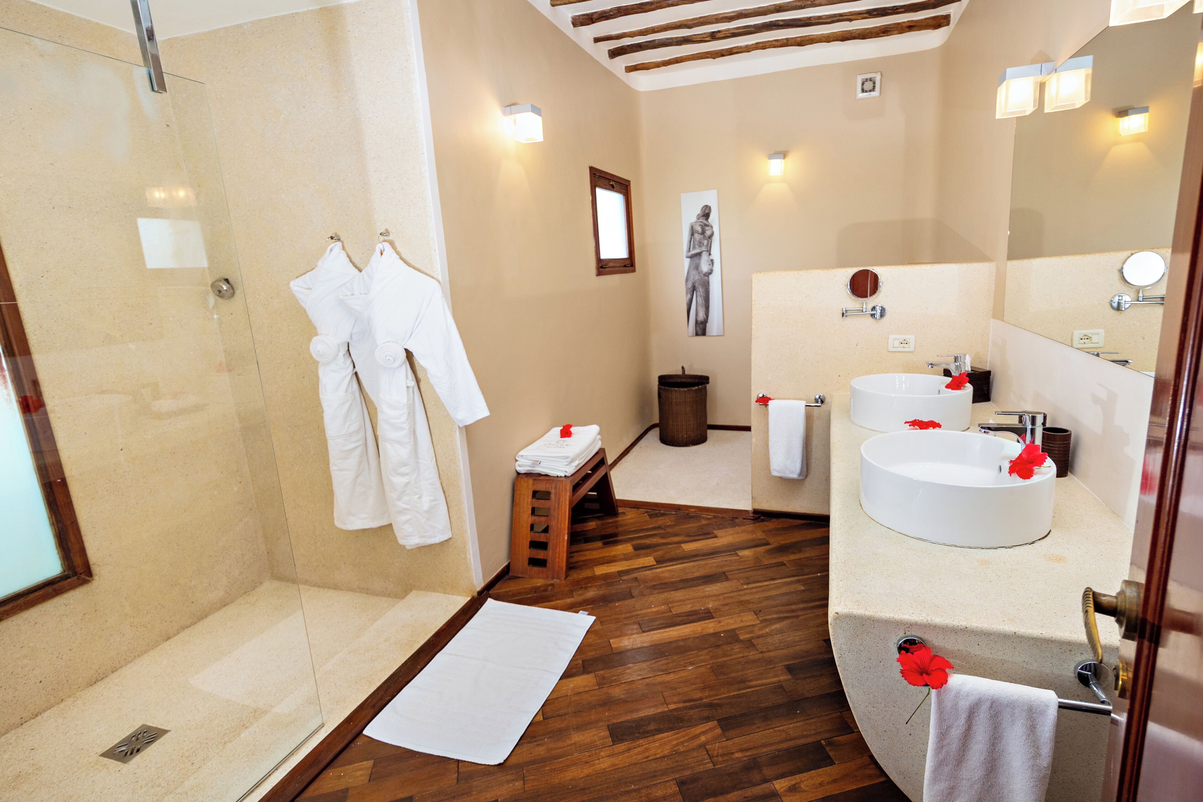 A bathroom with a glass shower enclosure, wooden flooring, a white sink with a round mirror, and a towel rack with white towels.