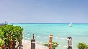 A wooden pier with rope railings leading to turquoise waters, a sailboat in the distance, and a clear sky. - Essque Zalu Zanzibar (Nungwi)