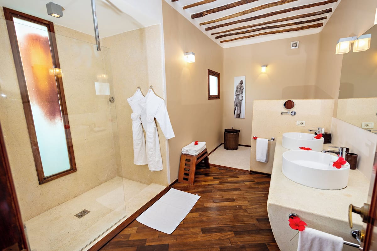 A bathroom with a glass shower enclosure, wooden flooring, a large mirror, and a white sink with red flowers.