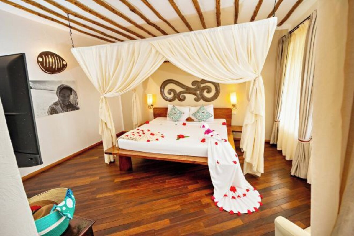 A bedroom with a canopy bed, wooden flooring, wall-mounted artwork, and a bed with rose petals.