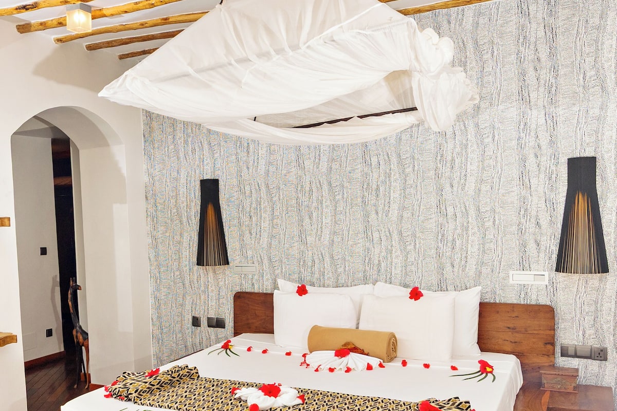 A room with a bed, wooden beams, a hanging canopy, a patterned wall, and a bench.