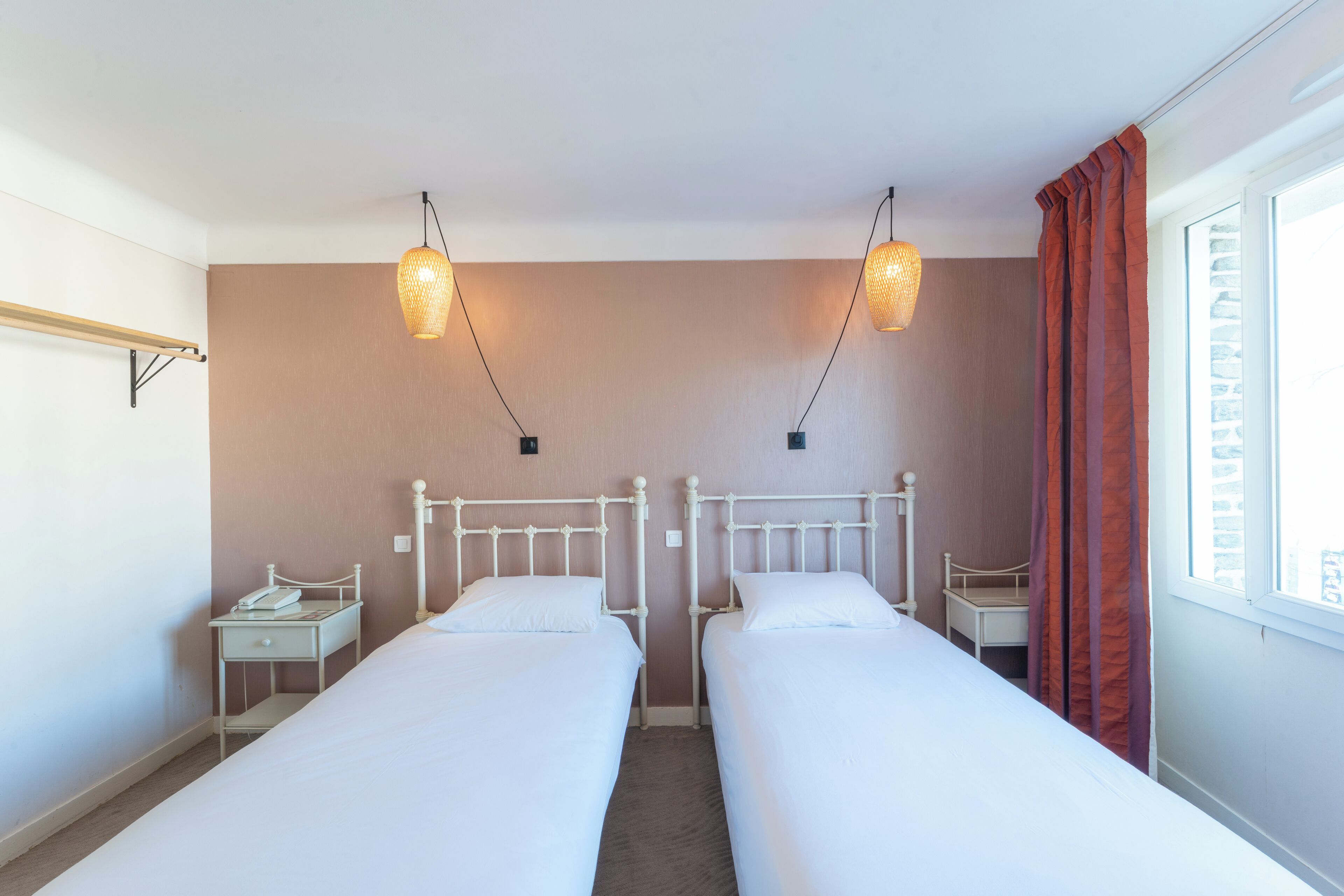 A twin room with two single beds, a small bedside table with a lamp, a window with curtains, and a wall-mounted shelf.