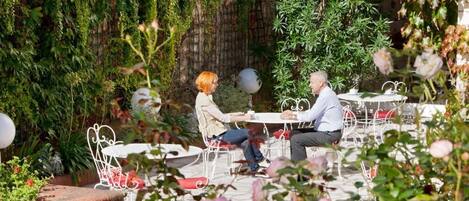 Un café al aire libre con mesas y sillas, rodeado de abundante vegetación y plantas.