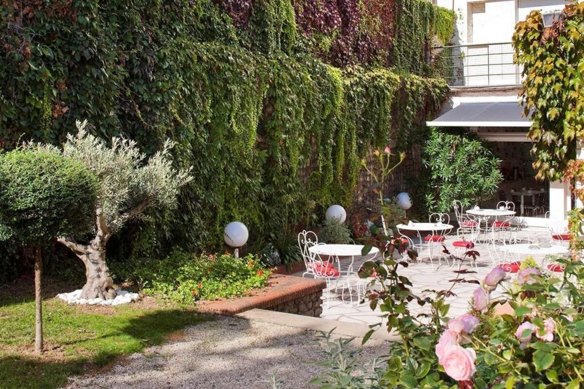 A garden patio with tables and chairs, surrounded by lush greenery and a tree.