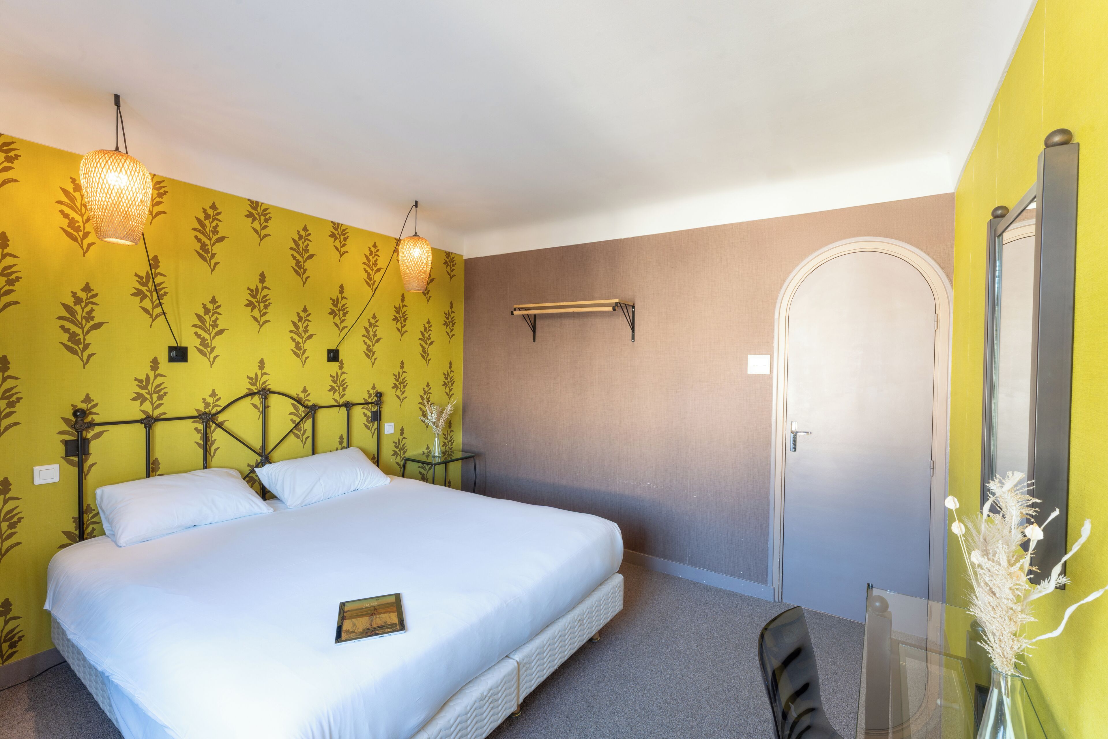 Una cama bien tendida en una habitación con paredes amarillas y marrones, una mesita con un florero y una puerta.