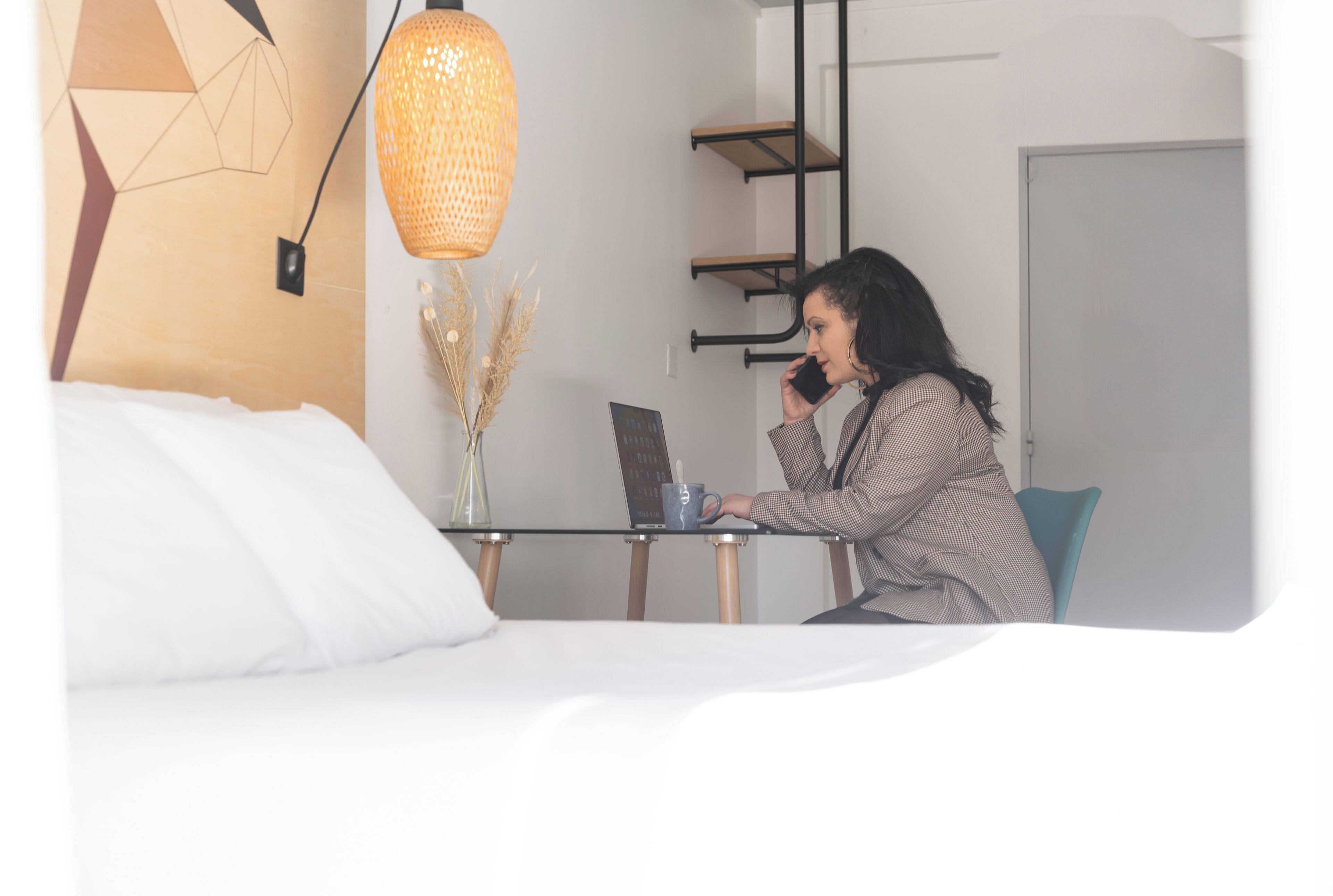 Una habitación con cama, escritorio, computadora portátil, taza azul y lámpara colgante.