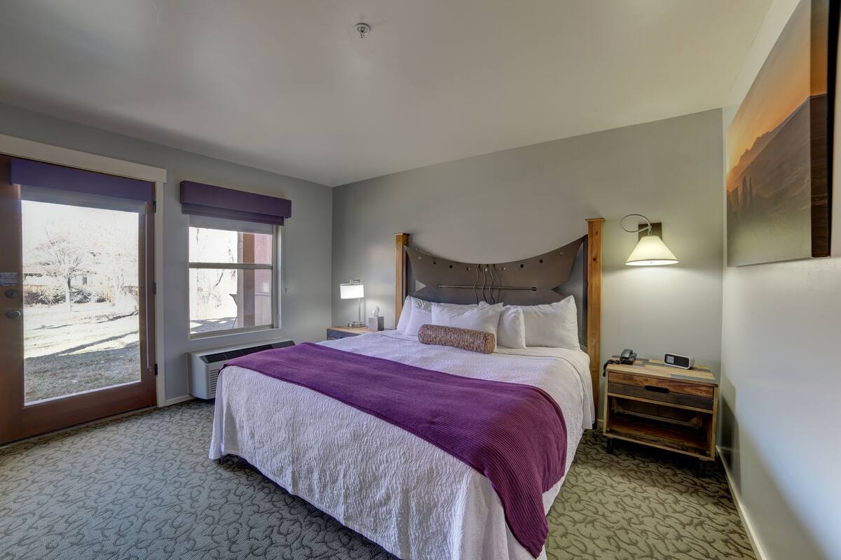 A hotel room with a large bed, a nightstand, a lamp, and a view of the outdoors through a sliding door.