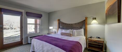 A hotel room with a large bed, a nightstand, a lamp, and a view of the outdoors through a sliding door.