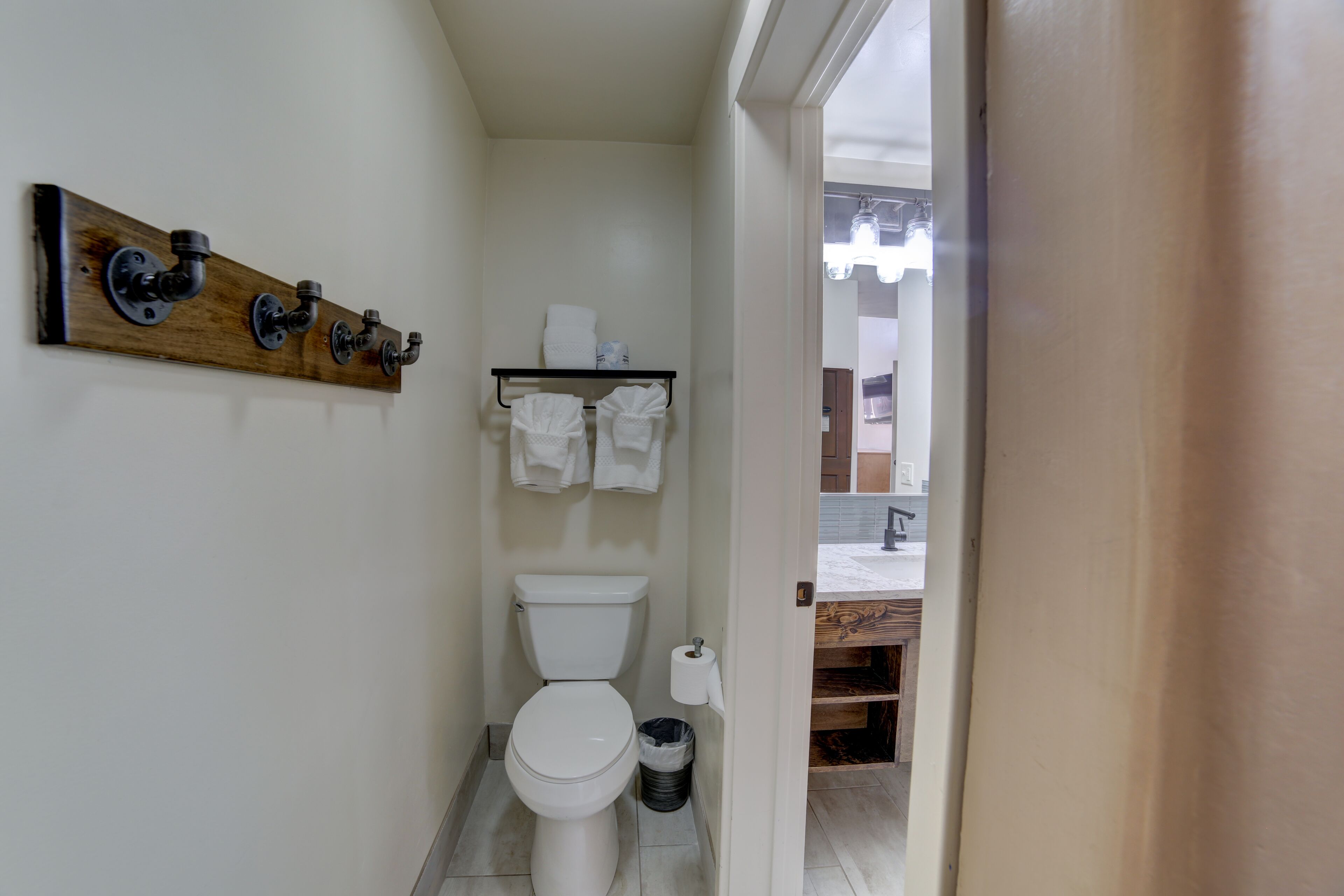 A compact bathroom with a white toilet, a wooden shelf with towels, and a black trash bin.