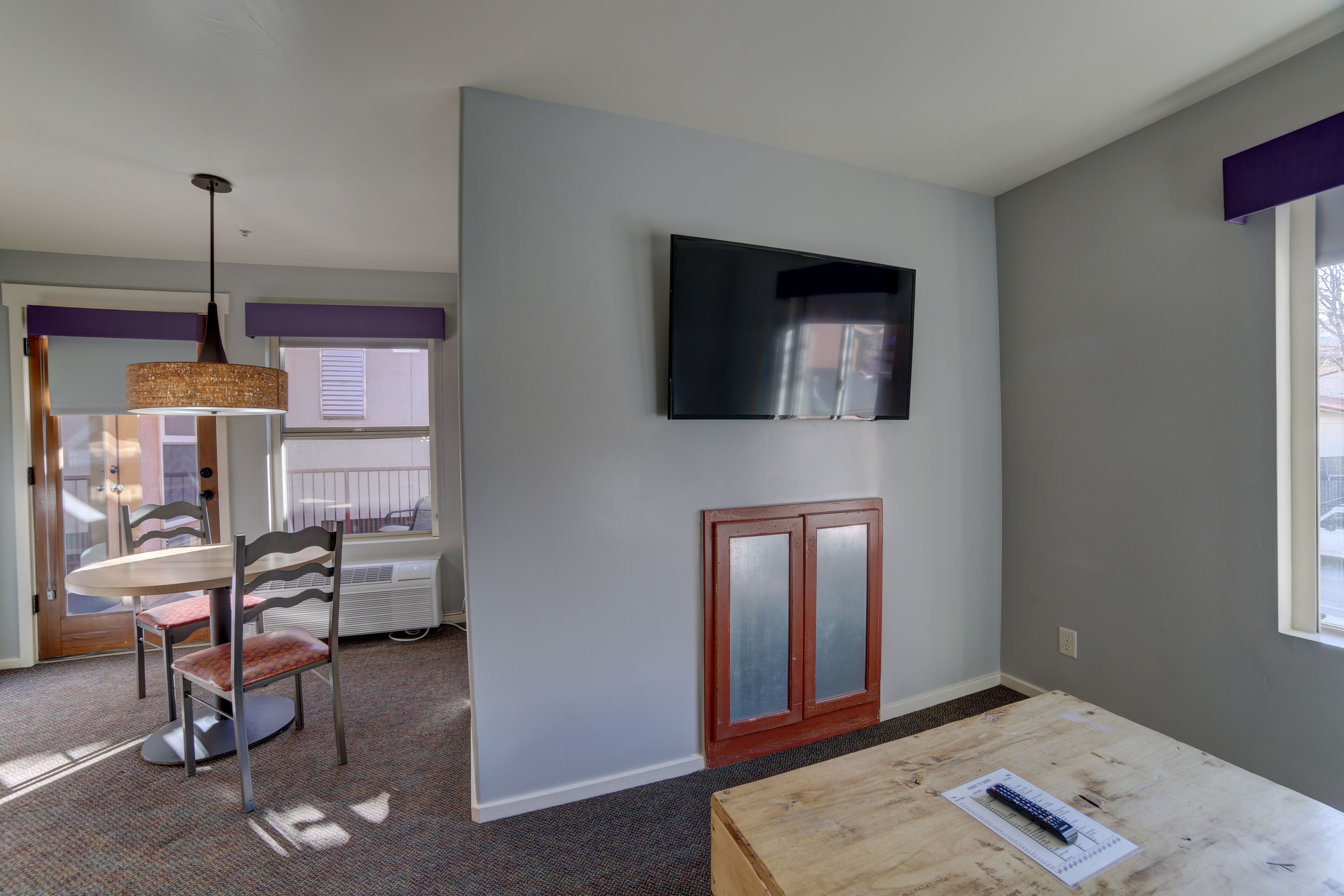 Una habitación con una mesa de comedor, sillas, un televisor montado en la pared y una ventana.