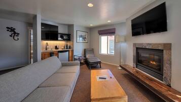 A modern living room with a sofa, a wooden coffee table, a fireplace, a TV mounted on the wall, and a kitchen area with a microwave and cabinets.