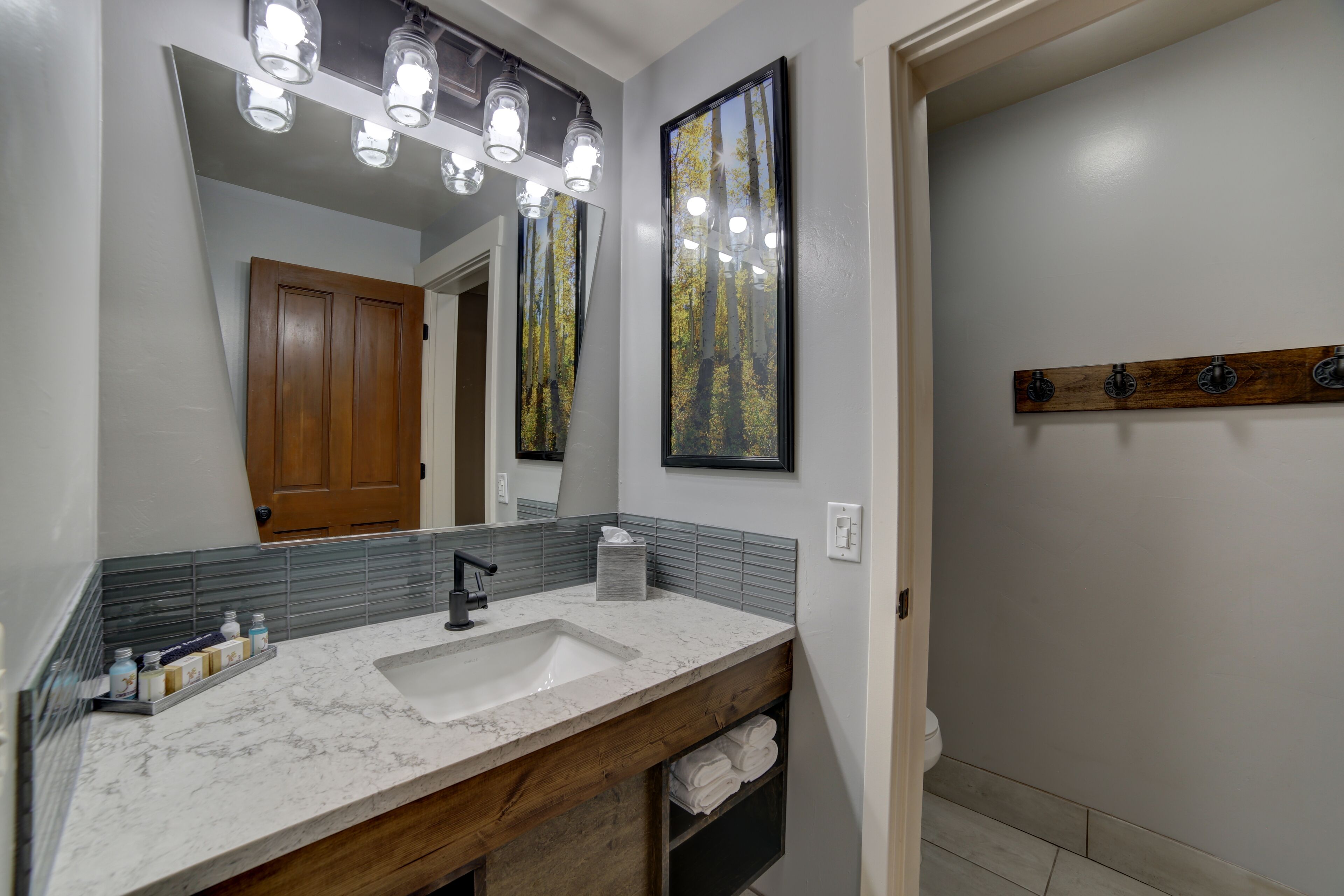 Une salle de bain avec un lavabo en marbre blanc, un meuble-lavabo en bois et un grand miroir agrémenté de trois lampes. Des tableaux encadrés ornent le mur, et un porte-manteau en bois est également présent.