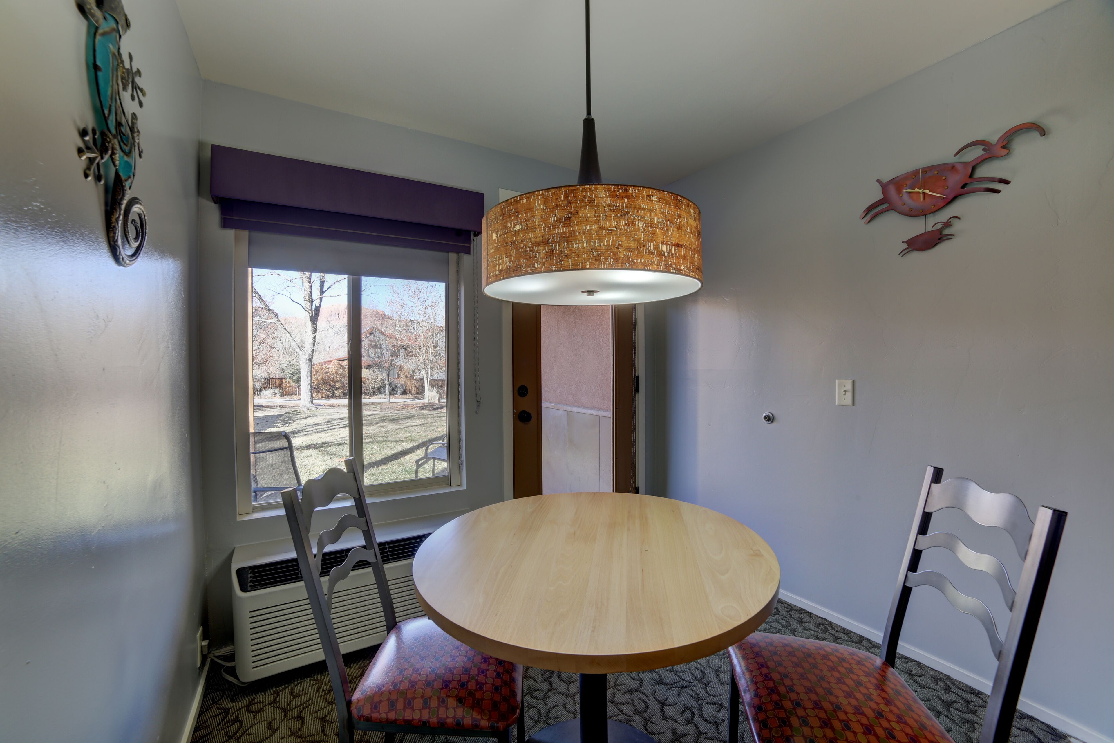 A dining area with a round wooden table, two chairs, a hanging light fixture, a window with a view, and wall art.