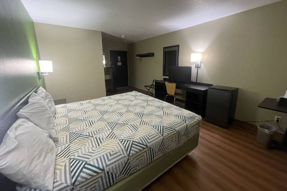 A hotel room with a bed, a desk, a chair, a television, and a trash bin.