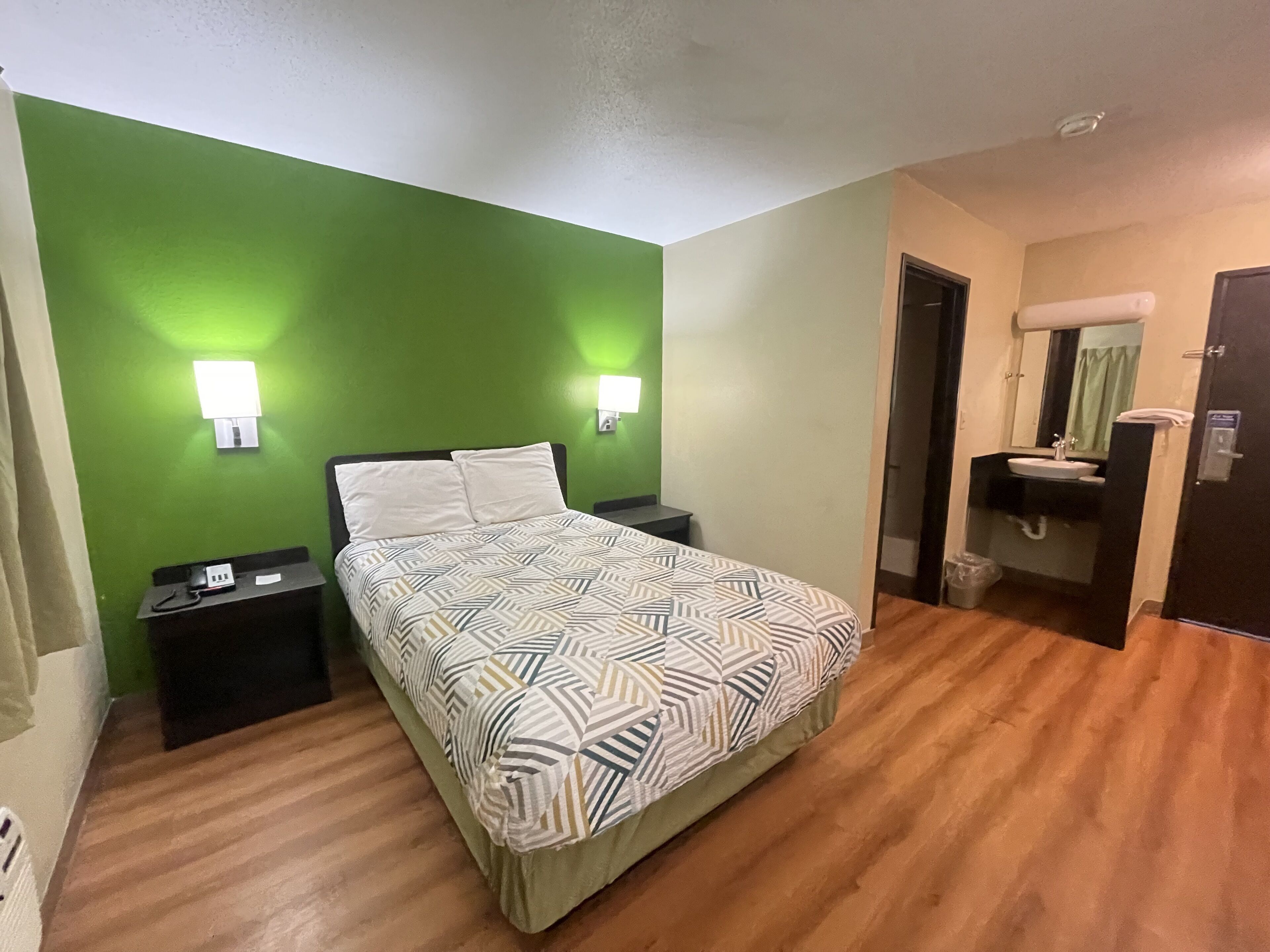 A hotel room with a bed, bedside tables, a green wall, a bathroom with a sink and mirror, and a door.
