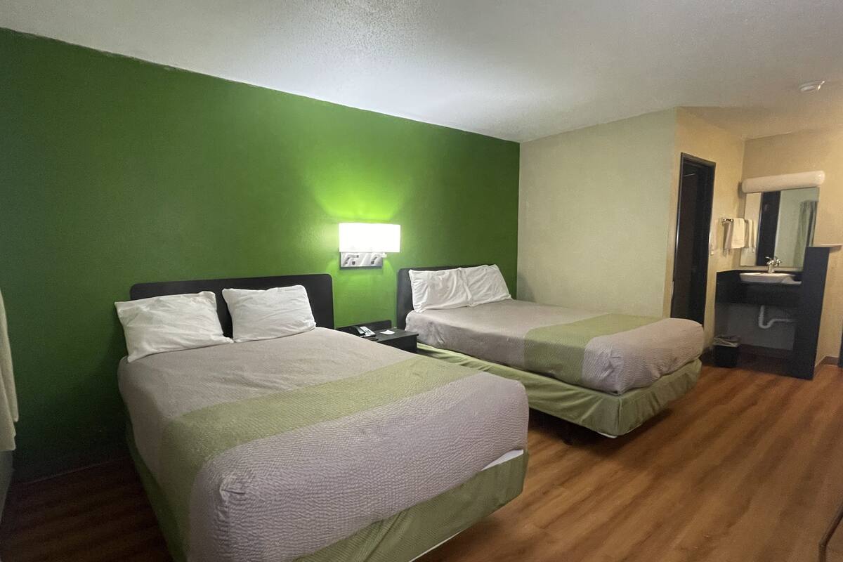 A hotel room with two beds, a nightstand, a lamp, and a bathroom with a sink and mirror.