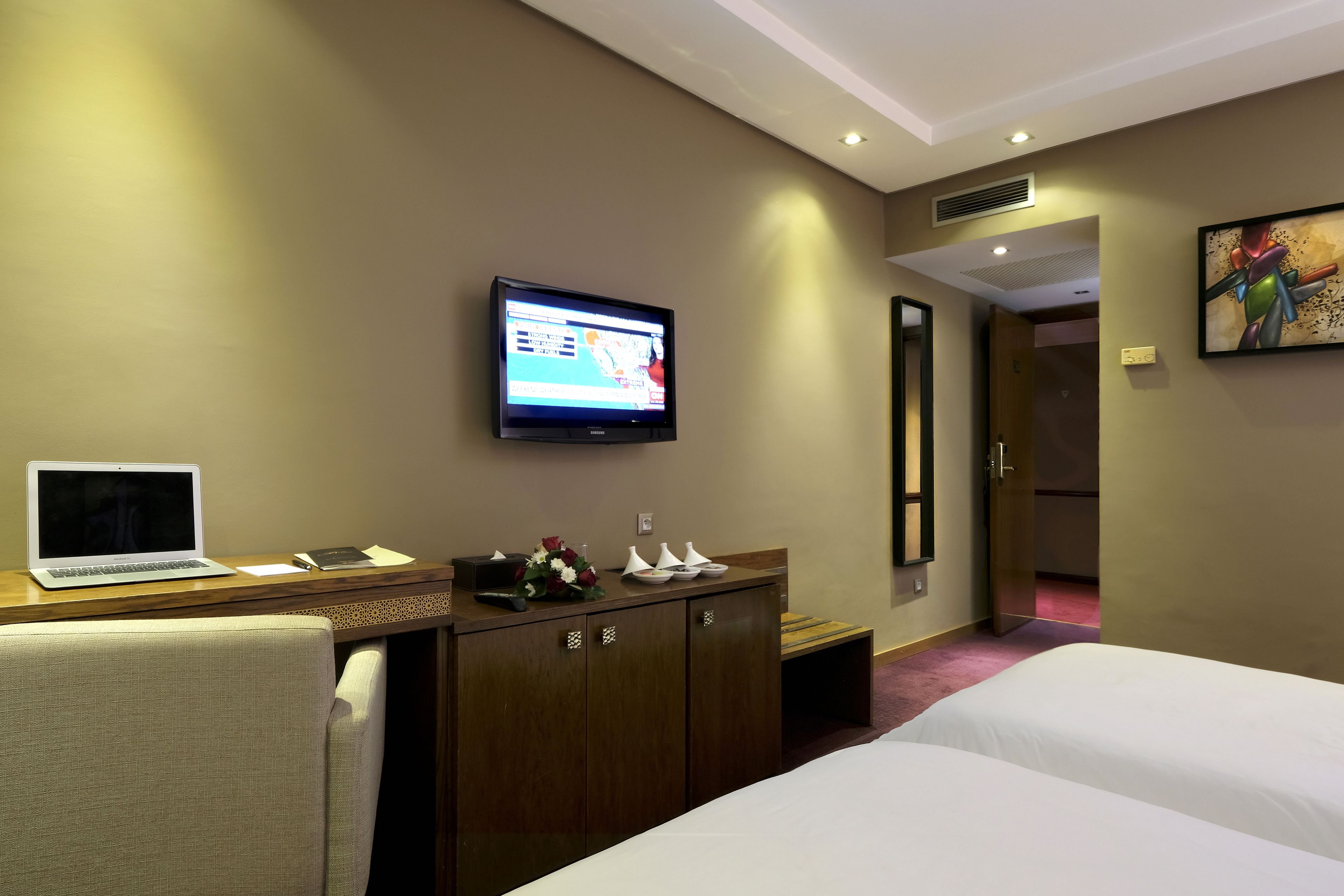 A hotel room with a bed, a desk with a laptop, a television, and a painting on the wall.