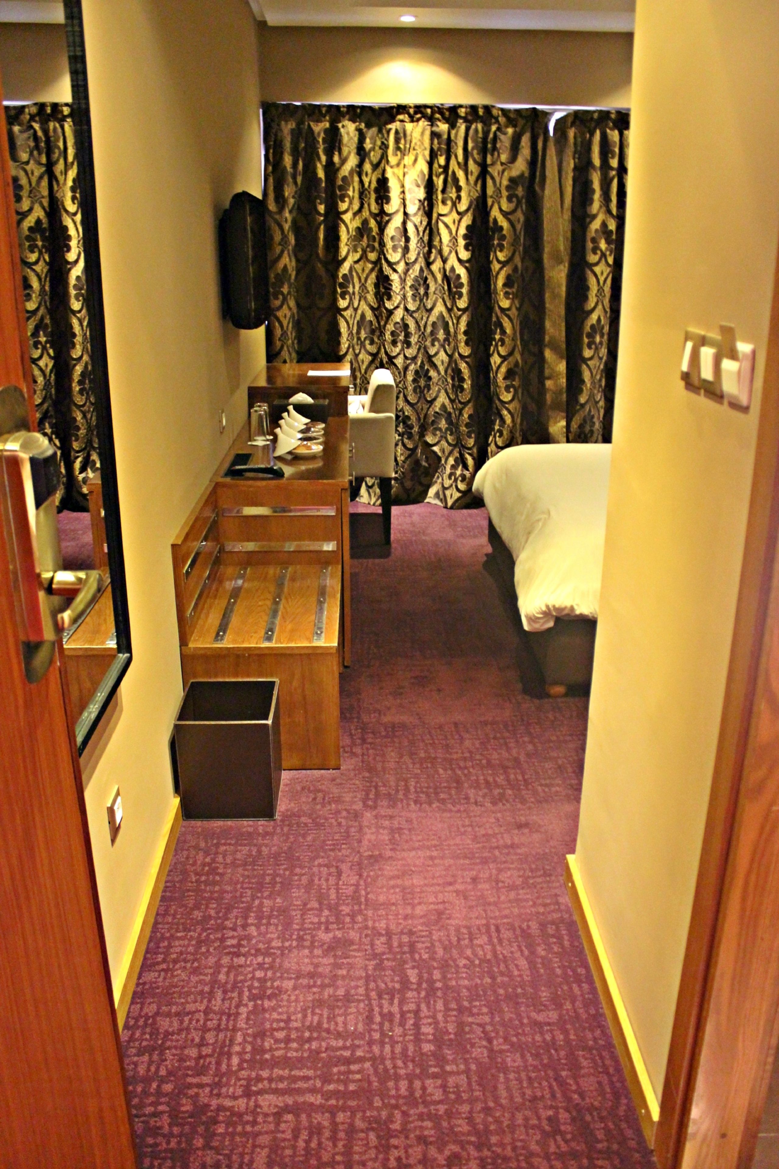 Une chambre d’hôtel avec un lit, un bureau, une chaise, un miroir, une petite poubelle et un rideau.