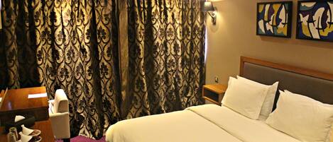 A hotel room with a large bed, a desk with a chair, a table with a lamp, and two paintings on the wall.