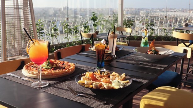 A dining table with a pizza, a salad, and a drink, set against a city view. - Hotel Belere Rabat (Rabat)