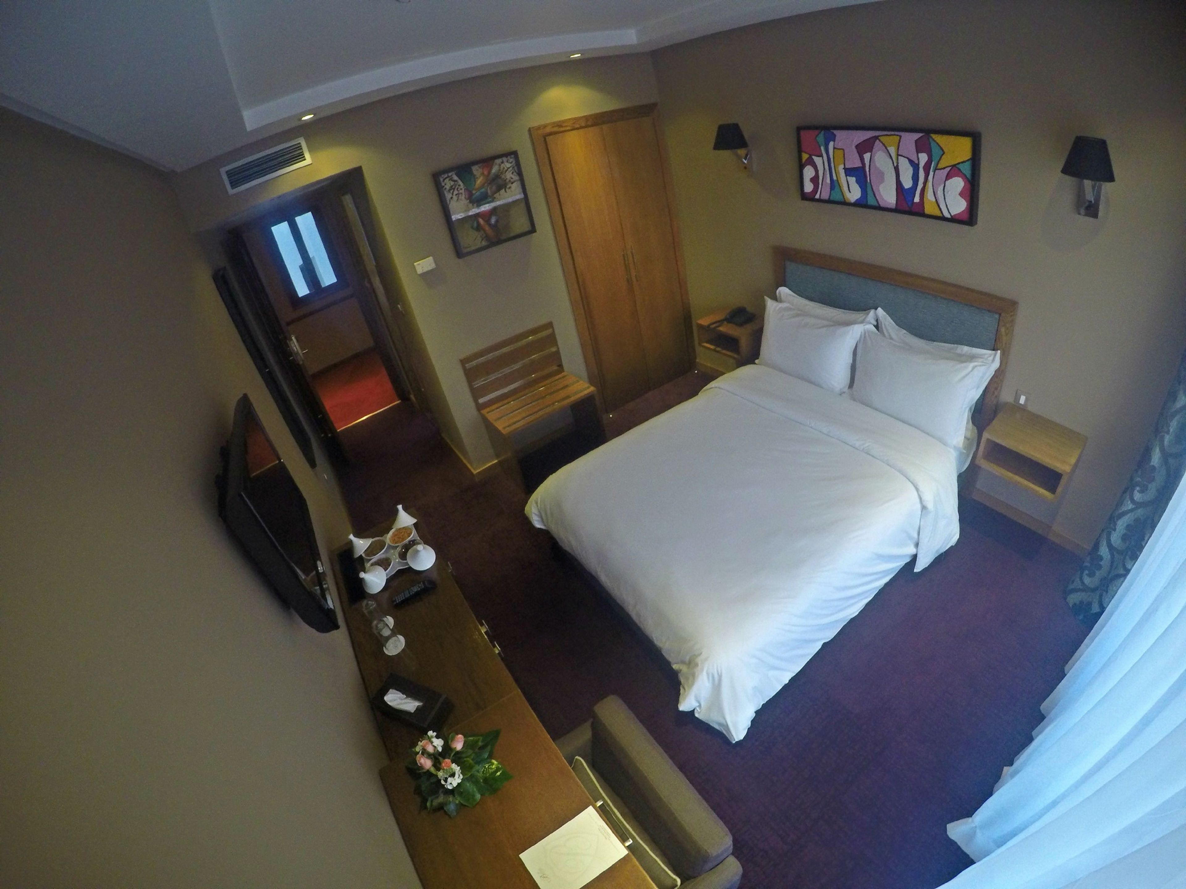 Habitación de hotel con cama, mesitas de noche, escritorio y silla, espejo, televisor y obras de arte en las paredes.
