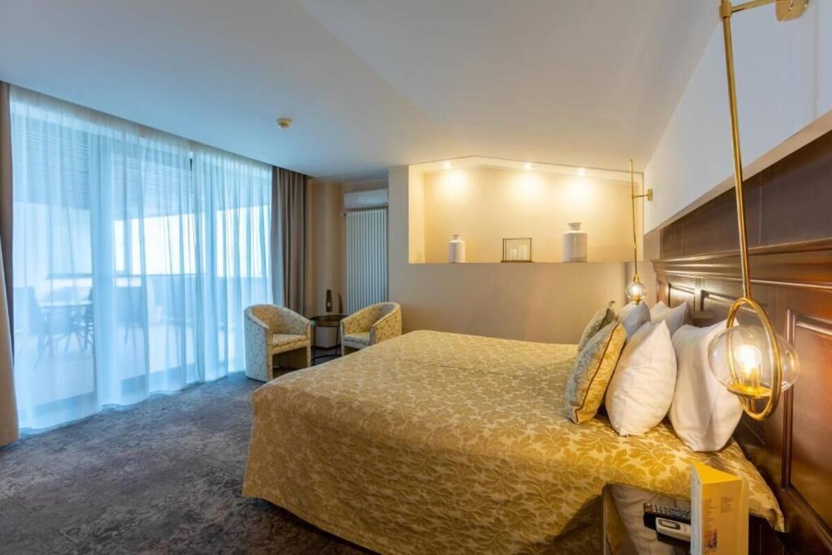 A hotel room with a large bed, a sitting area with a chair, a small table, and a large window with curtains.