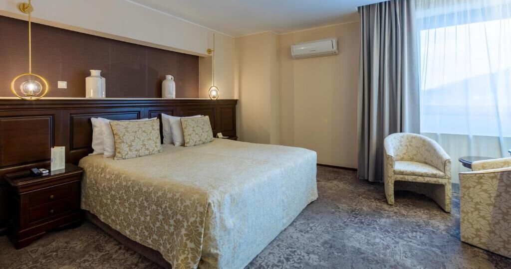 Habitación de hotel con una cama grande, una mesita de noche, una silla, una pequeña mesa y un ventanal con cortinas.