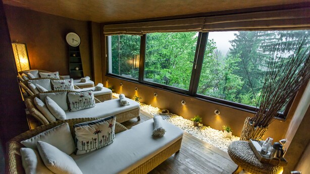 A room with a large window, a sofa with cushions, a clock on the wall, and a small table with decorative items.