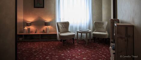 A room with two chairs, a small table, a lamp, a picture on the wall, and a window with sheer curtains.