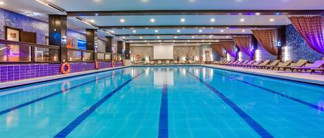 A large indoor swimming pool with clear blue water, surrounded by lounge chairs and a tiled wall with a life buoy.