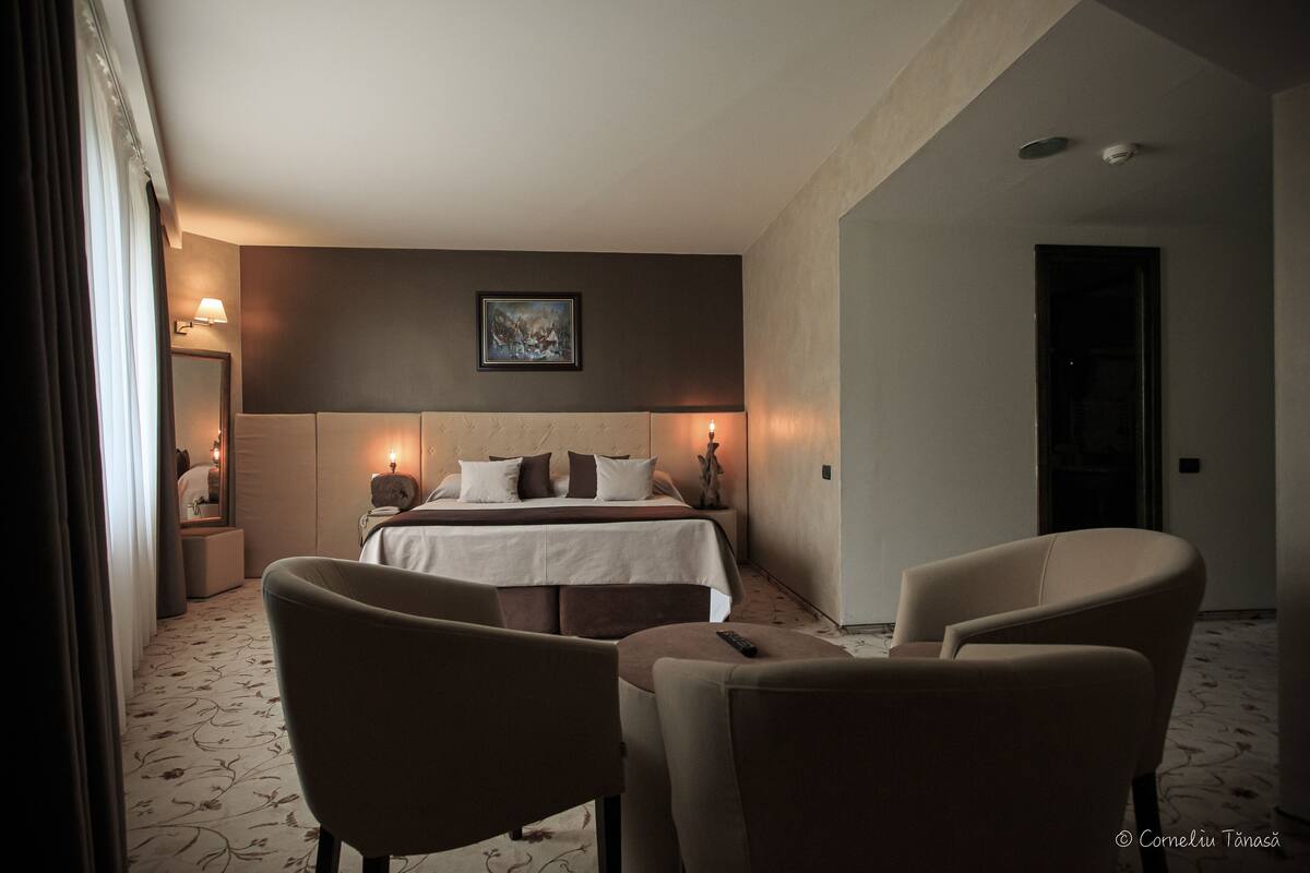 A hotel room with a bed, two chairs, a nightstand, and a painting on the wall.