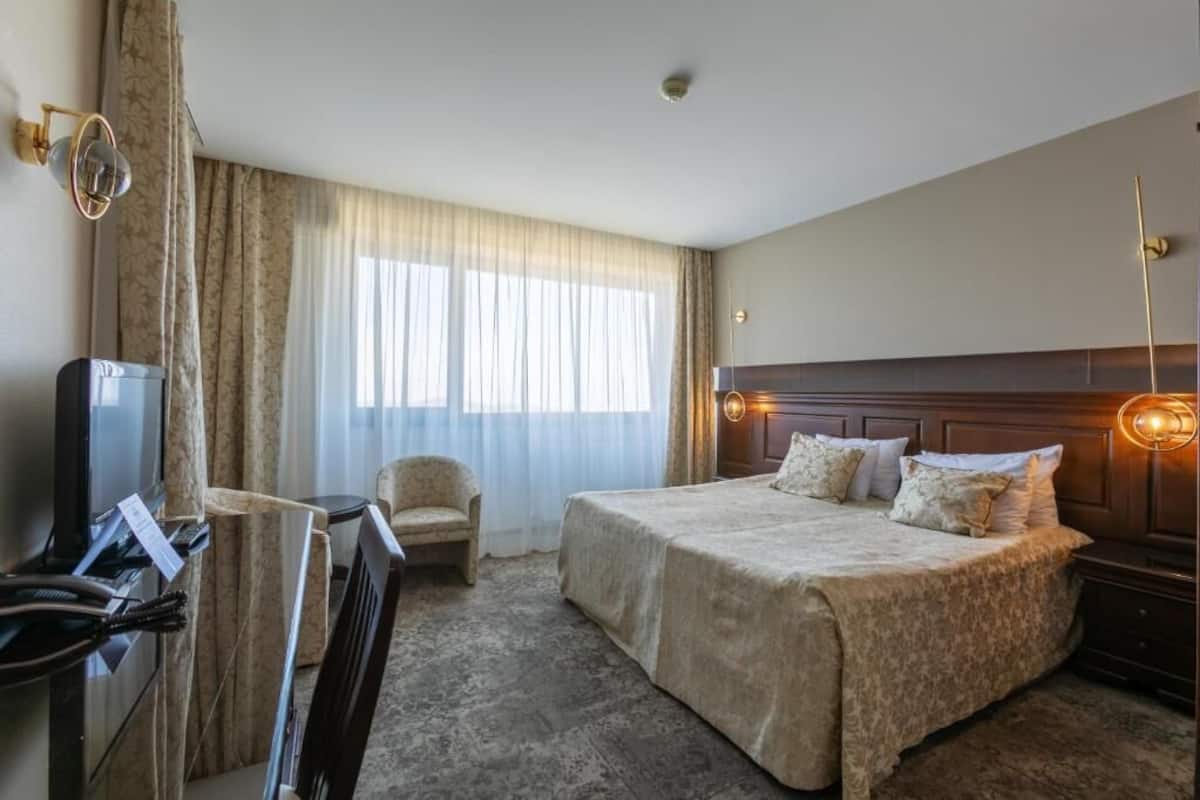 A hotel room with a large bed, a desk with a television, a chair, and a window with curtains.