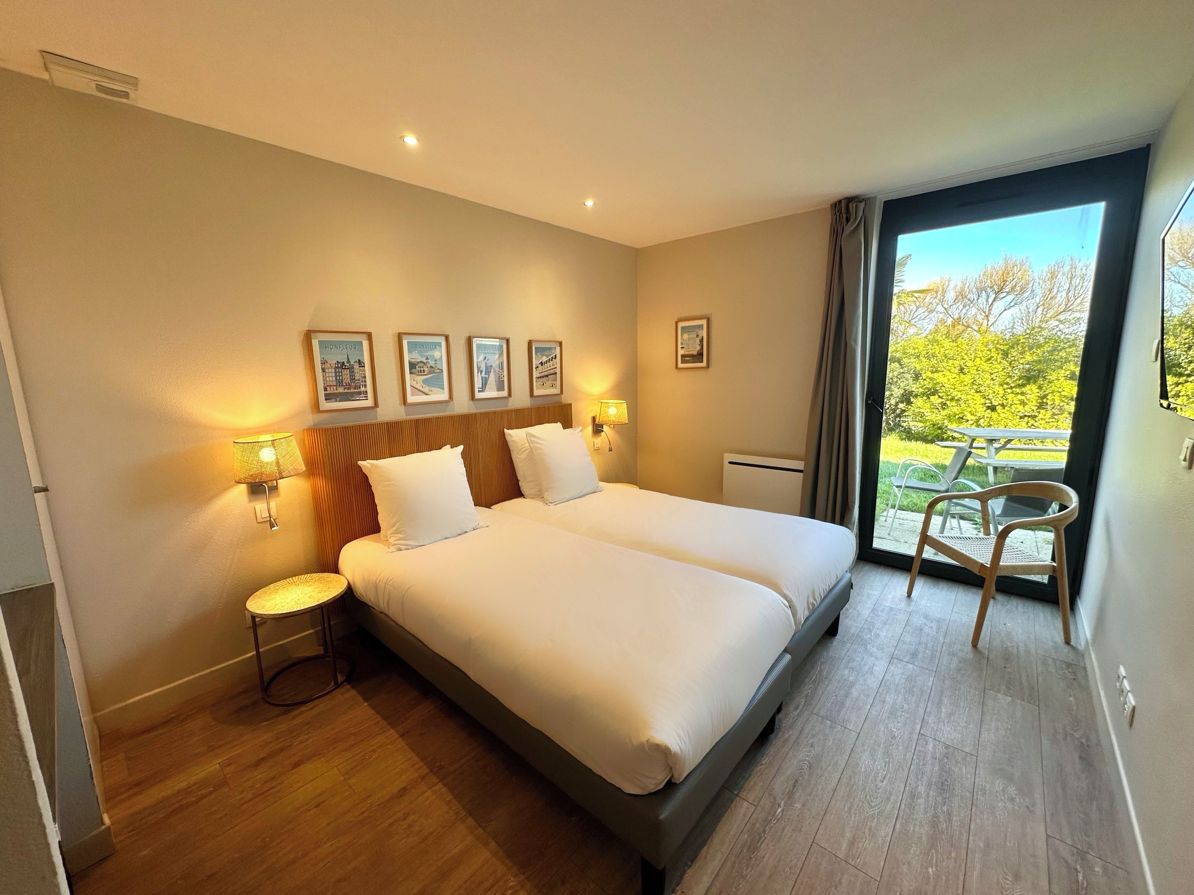 A hotel room with a double bed, two bedside lamps, a small table, a chair, and a balcony with a table and chairs.
