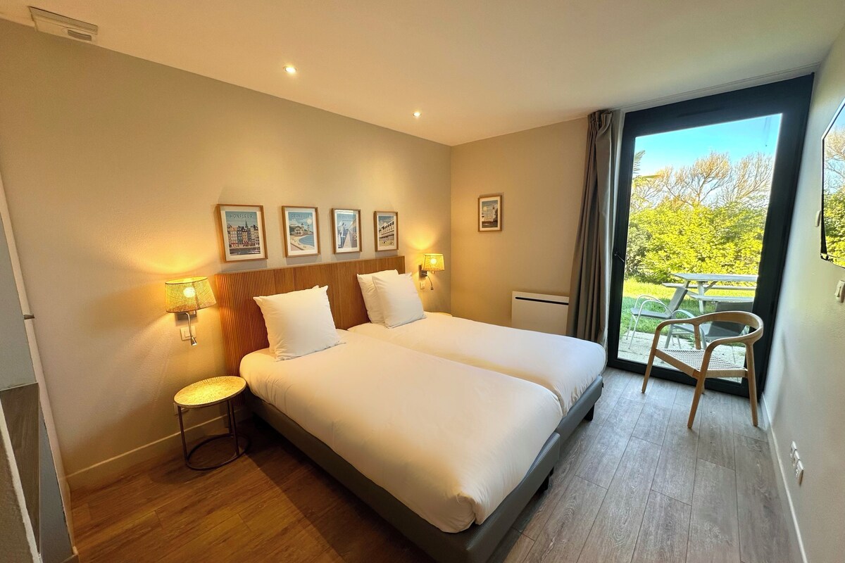 Habitación de hotel con cama doble, dos lámparas de noche, una mesita, una silla y un balcón con mesa y sillas.