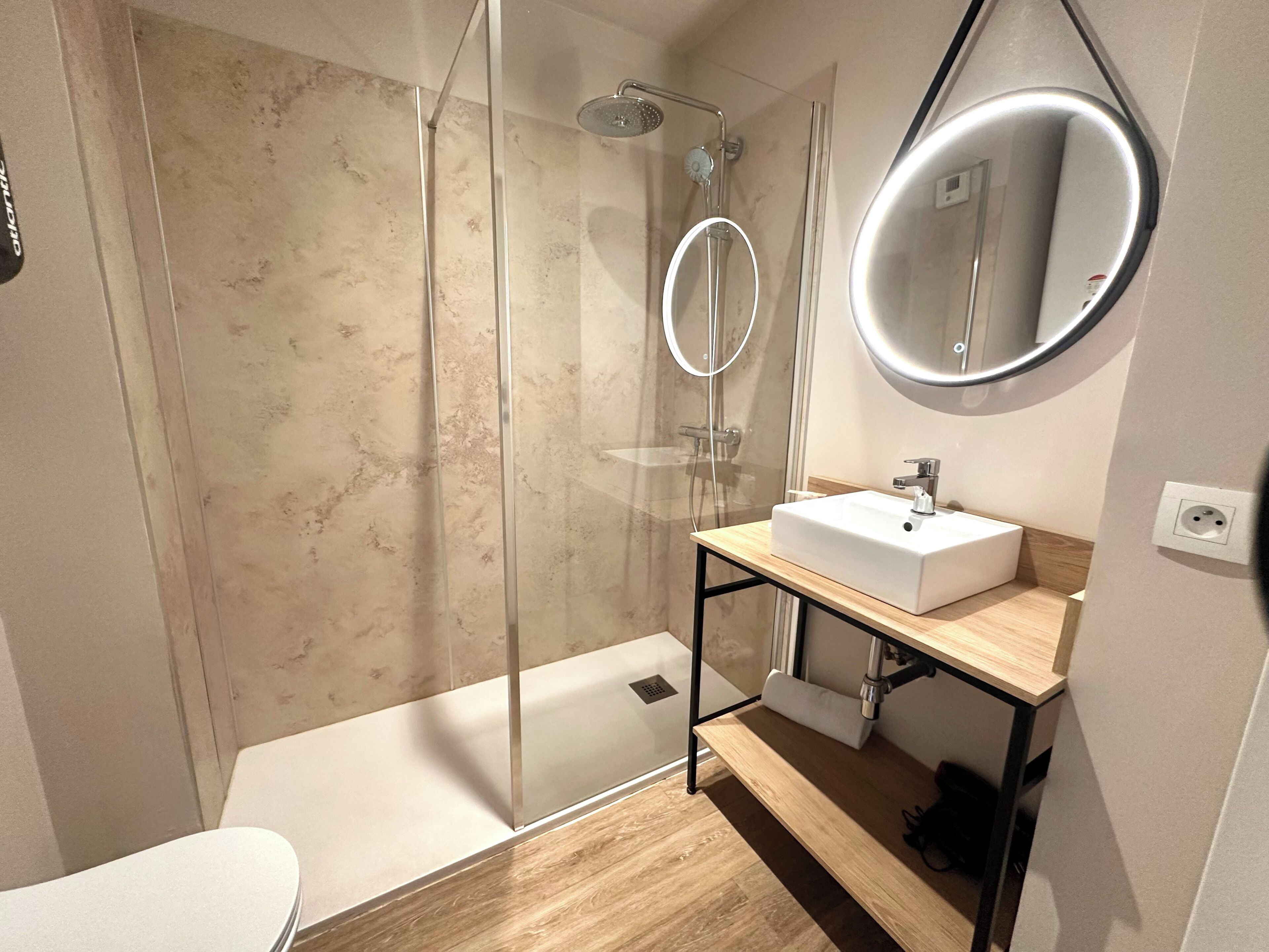 Un bany modern amb dutxa de vidre, un lavabo blanc i un mirall rodó.