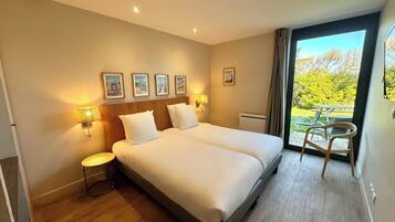 A hotel room with a double bed, two bedside lamps, a small table, a chair, and a balcony with a table and chairs.