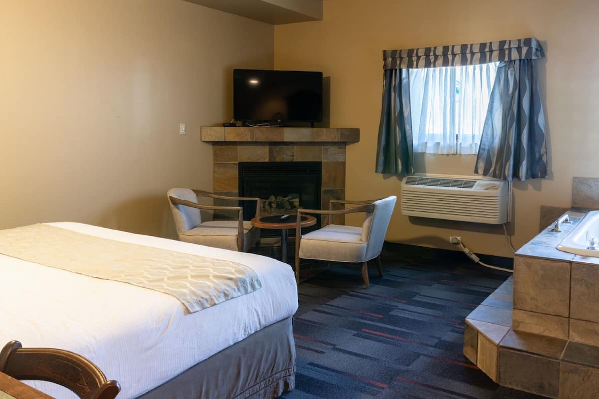 A hotel room with a bed, a seating area with chairs, a fireplace, a television, an air conditioning unit, and a bathtub.