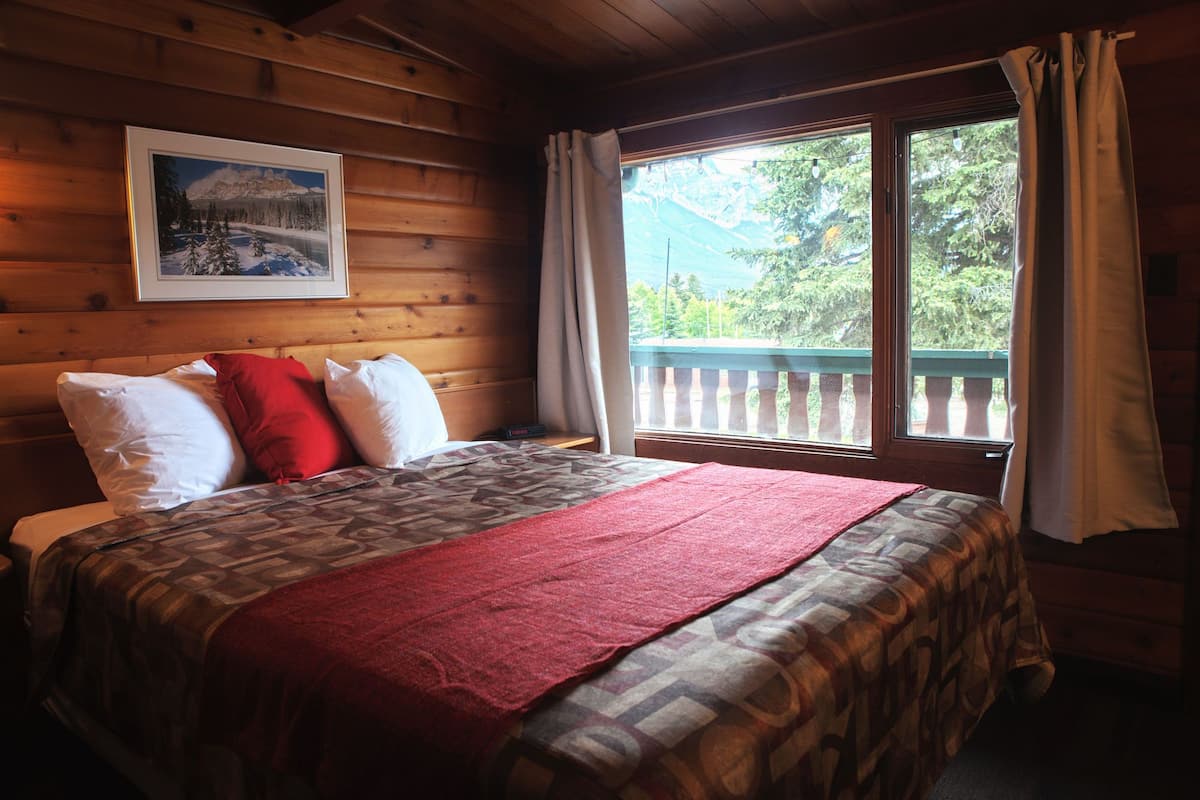 A wooden cabin room with a bed, a framed picture on the wall, and a window with a view of greenery.