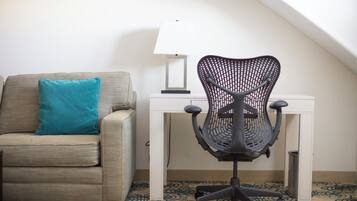 A room with a beige sofa, a teal pillow, a black office chair, a white desk, and a patterned carpet.