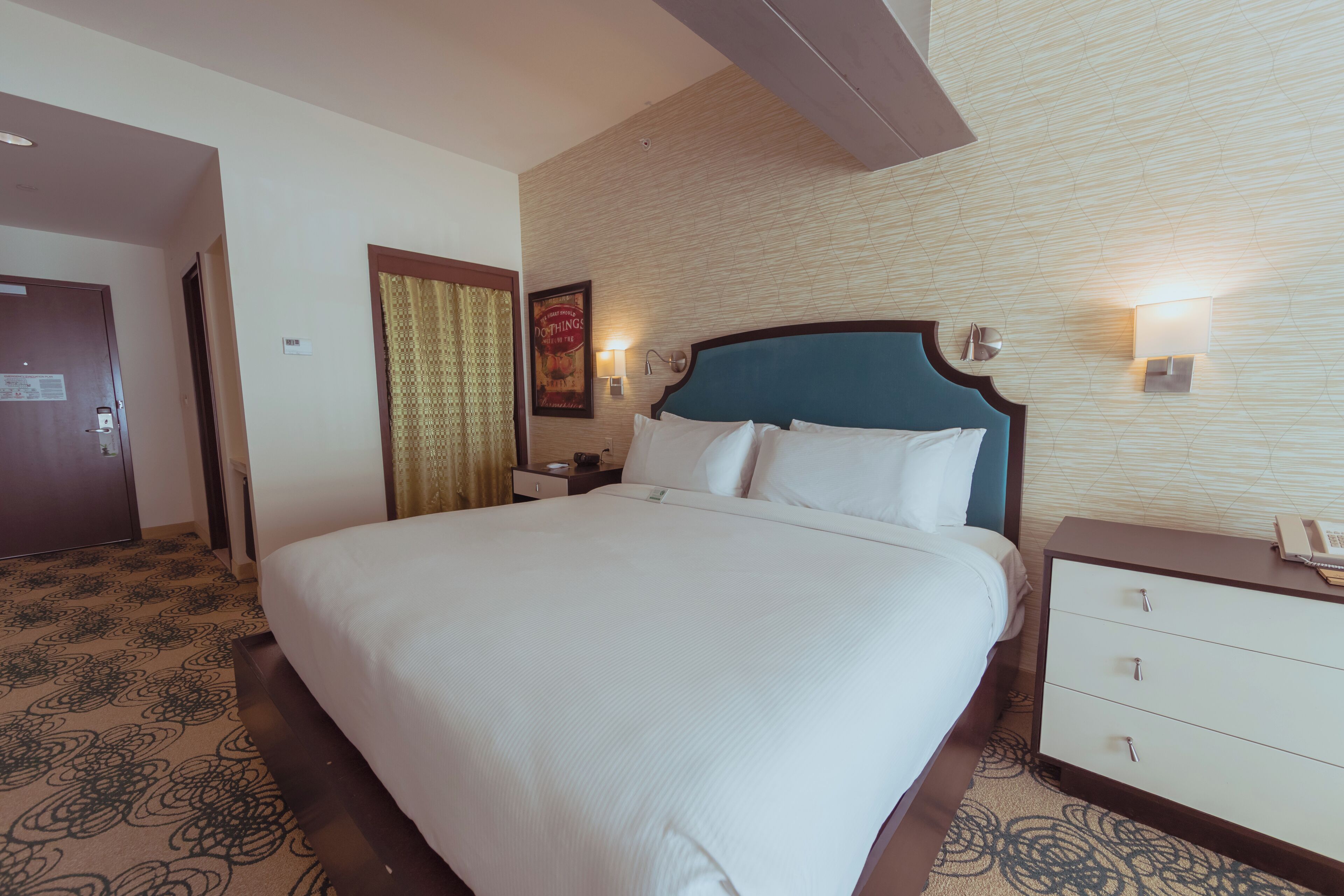 A hotel room with a large bed, a nightstand with a lamp, and a wall-mounted light.