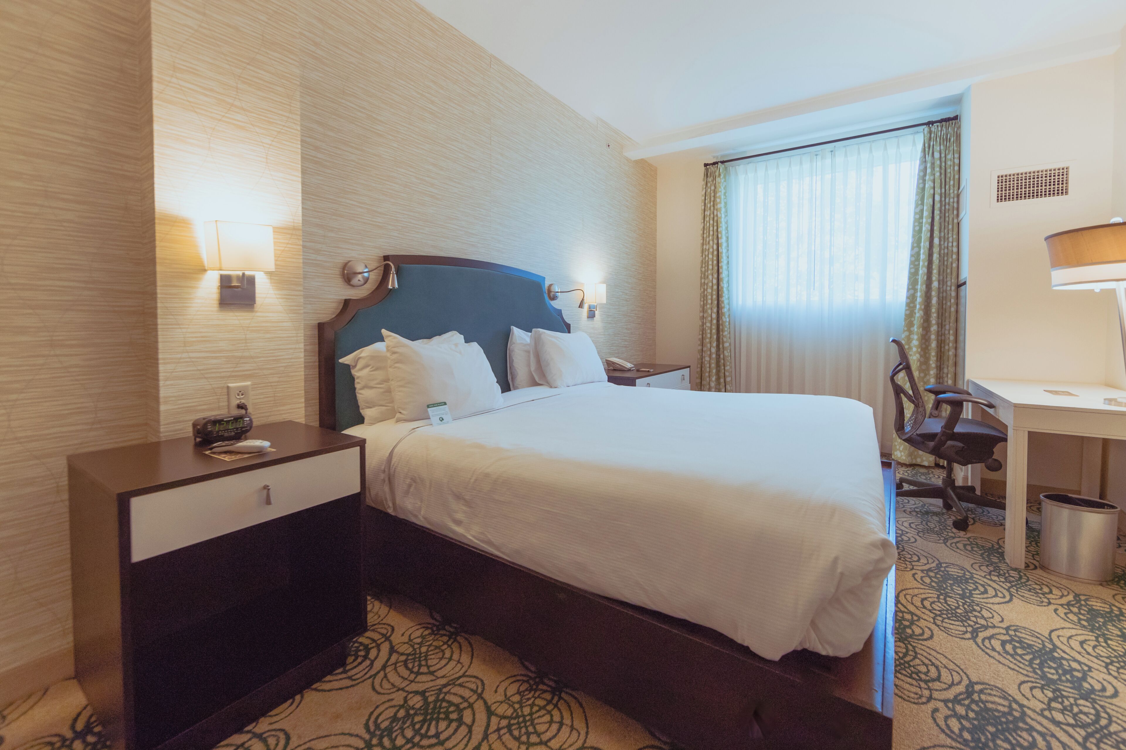 A hotel room with a large bed, a desk, a chair, a nightstand, and a lamp.