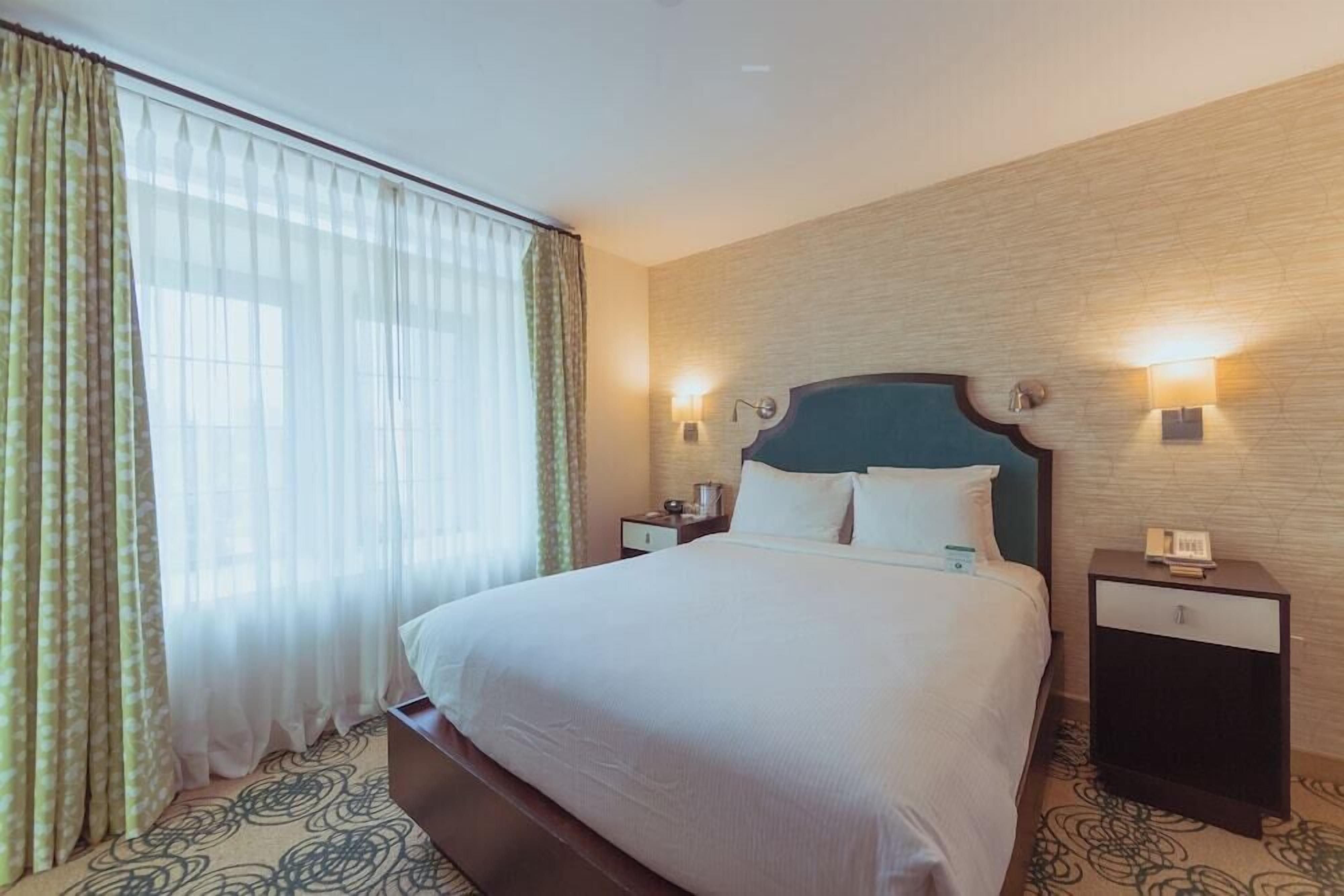 Quarto de hotel com uma cama grande, duas mesas de cabeceira, um criado-mudo e uma janela com cortinas.