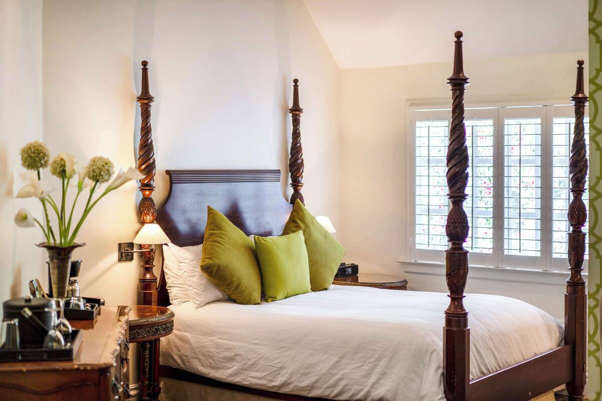 A four-poster bed with a headboard, green and white pillows, a bedside table with a lamp, and a window with a view of greenery.