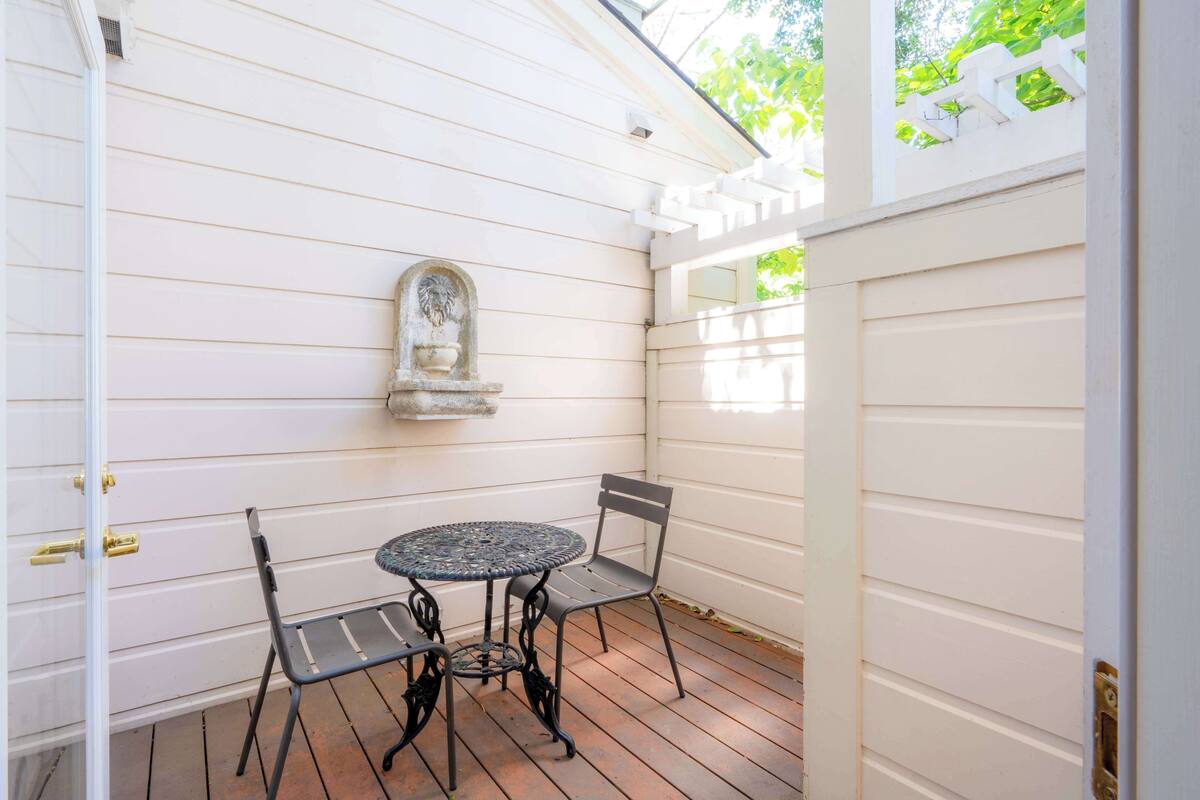 A covered patio with a round table and two chairs, a stone wall with a decorative niche, and a white fence.
