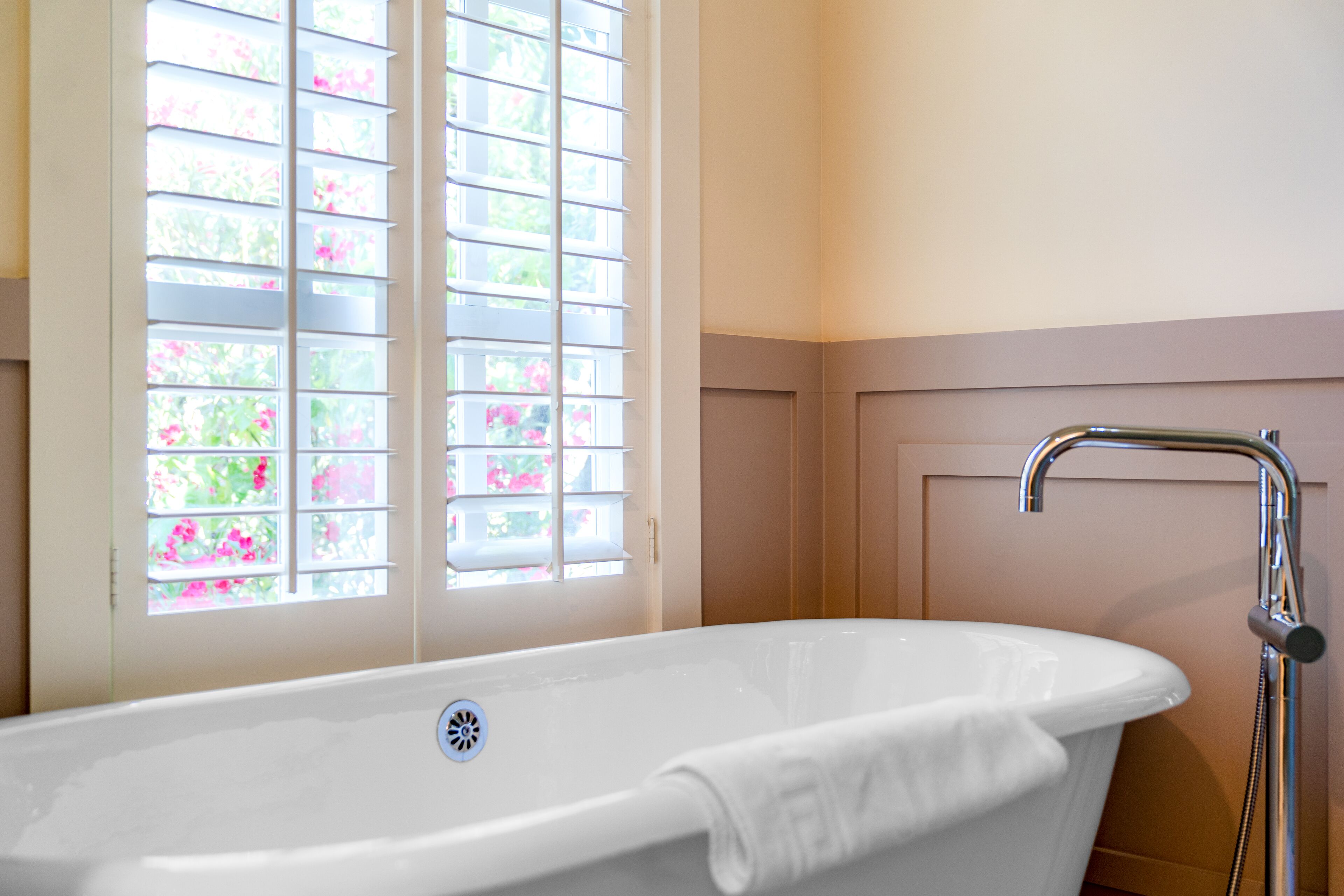 A white freestanding bathtub with a showerhead, a window with blinds, and a door with a handle.